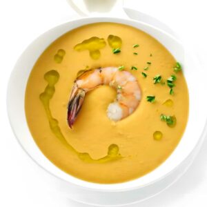Bowl of orange soup with shrimp, herbs, olive oil, and a spoon.