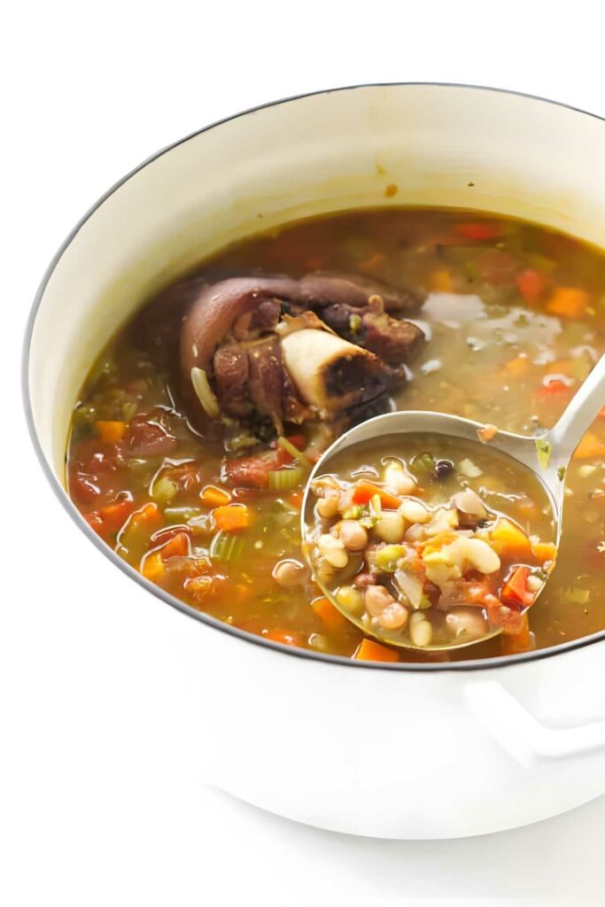 White pot of mixed bean soup with ham hock, ladle lifting portion.