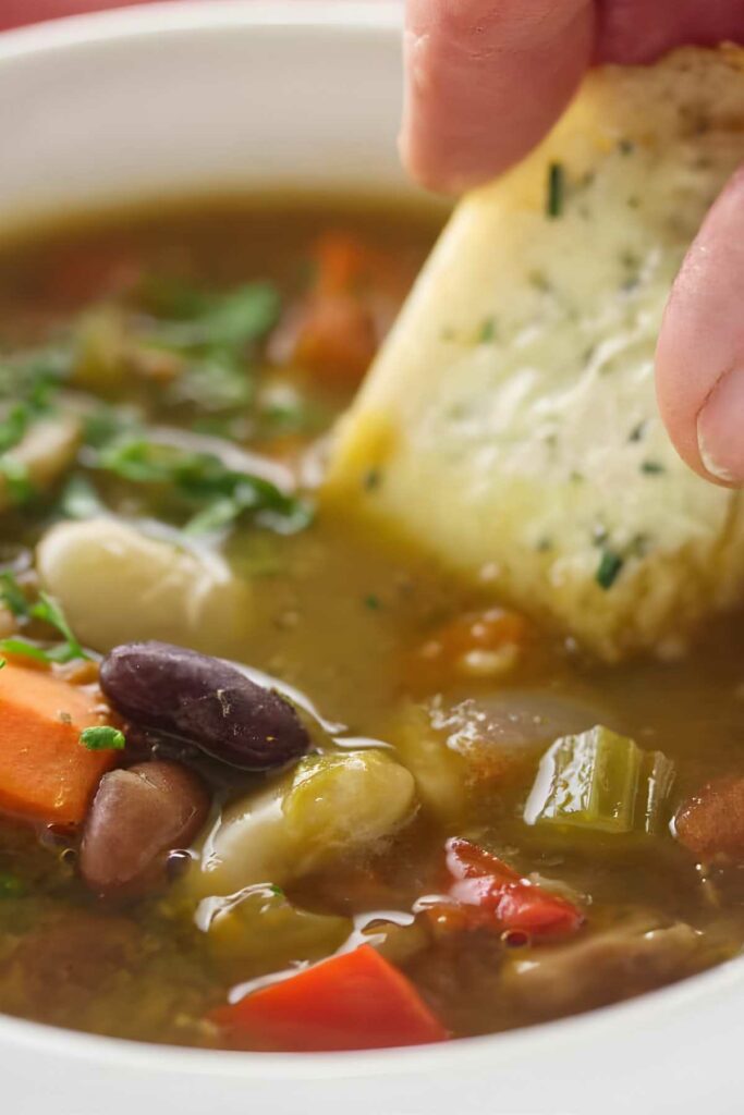 Hand dipping garlic bread into vegetable and bean soup with visible veggies.