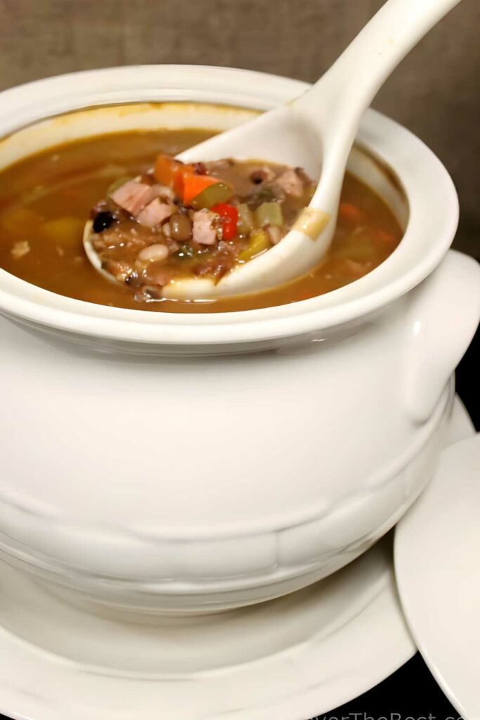 White ceramic tureen of soup with beans, vegetables, meat, and ladle.