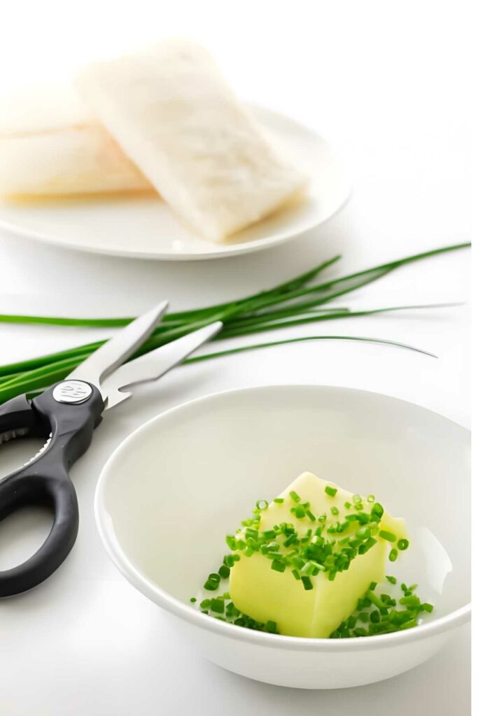 Bowl of butter with chives beside scissors; raw fish in background.