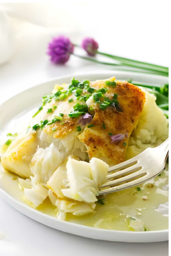 Fork breaking broiled white fish with chives, light sauce, on white plate.