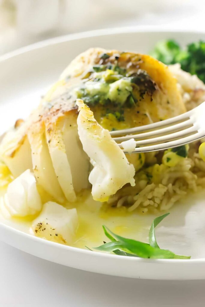 Fork cutting into baked white fish with lemon herb sauce on rice and vegetables.