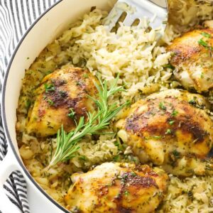 Baked chicken thighs on seasoned rice in a white pot, garnished with herbs.