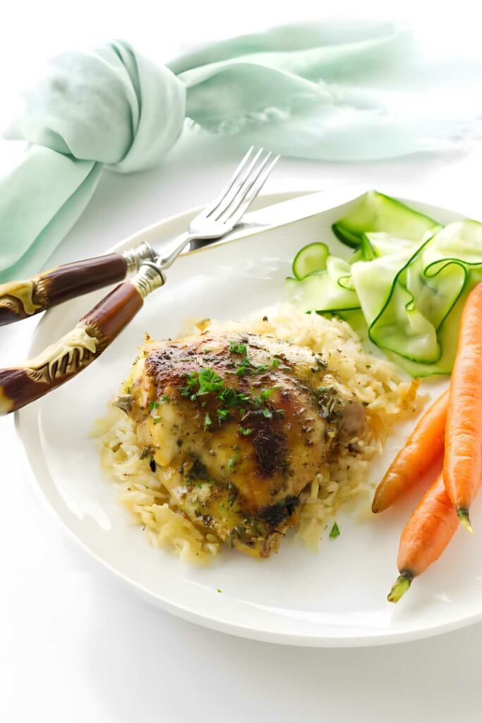 Baked chicken on rice with carrots, cucumber ribbons, herbs, and utensils nearby.