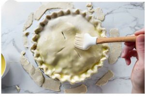 Hand brushing egg wash on crimped pie; dough scraps on white marble.