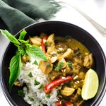 Black bowl with rice, yellow curry chicken, red peppers, herbs, and peanuts.