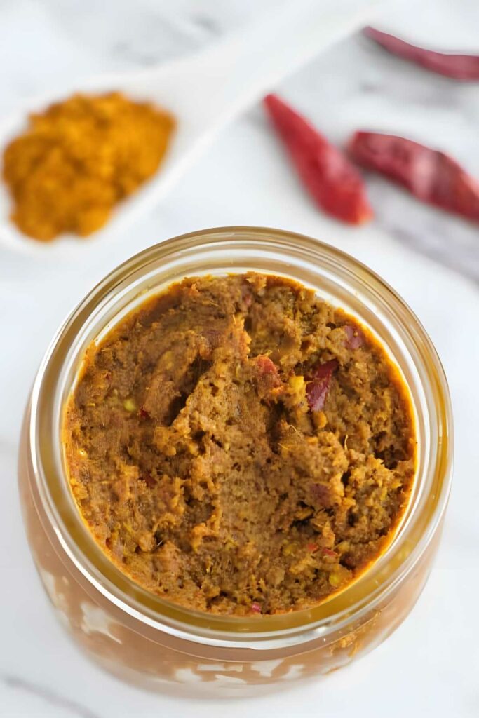 Jar of chunky brown curry paste with chilies and yellow curry powder behind.