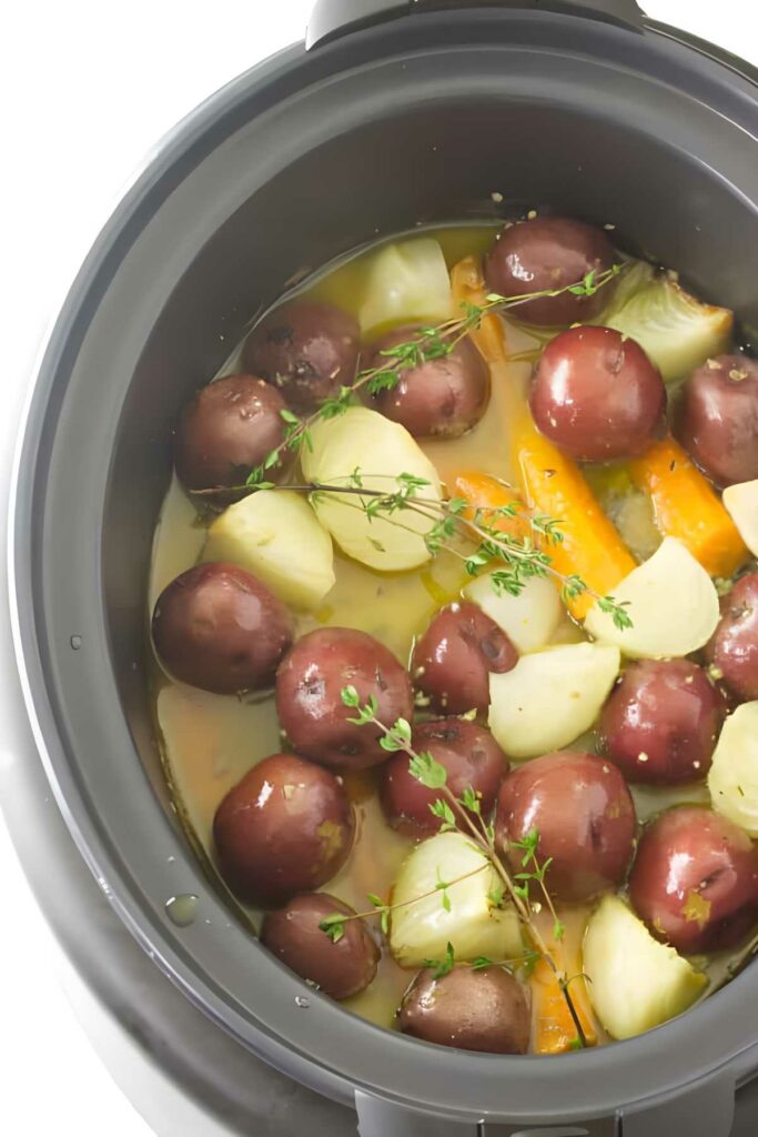 Slow cooker with red potatoes, carrots, onions, and thyme in broth.