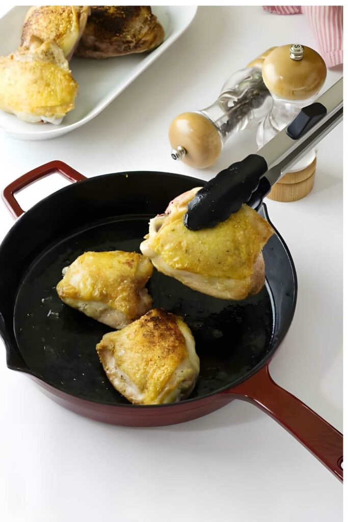 Tongs lifting cooked chicken over skillet with two pieces, pepper grinder behind.