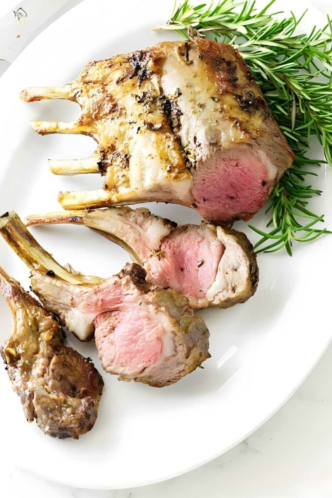 Sliced roasted rack of lamb on a white plate with rosemary garnish.