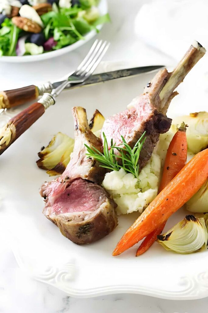 Two lamb chops on mashed potatoes with roasted vegetables and rosemary garnish.