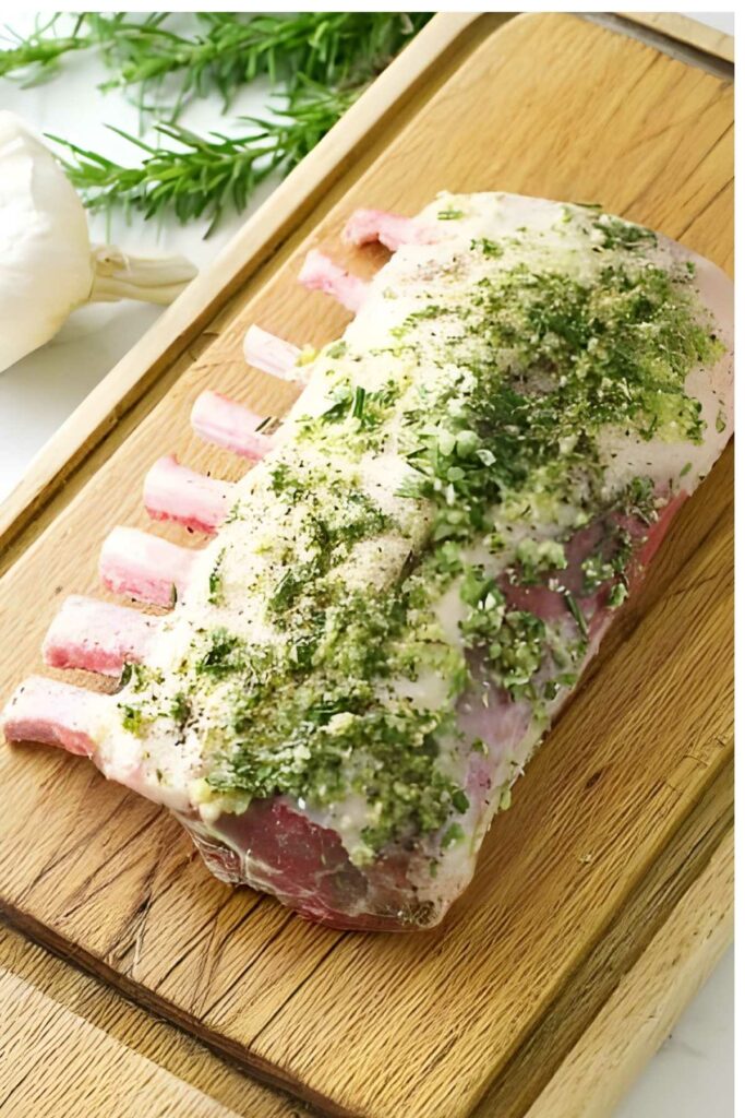 Herb-crusted raw rack of lamb on cutting board with rosemary and garlic.