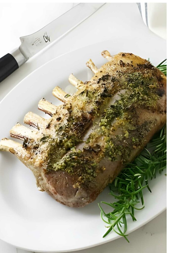 Herb-crusted rack of lamb on white plate, garnished with rosemary. Chef&rsquo;s knife nearby.