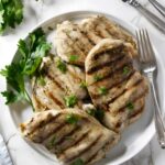 Four grilled chicken breasts with parsley on a white plate, utensils beside.