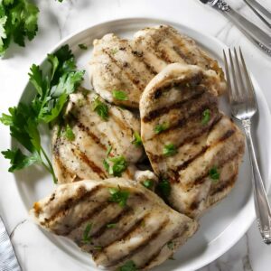 Four grilled chicken breasts with parsley on a white plate, utensils beside.
