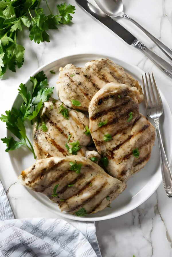 Four grilled chicken breasts with parsley on a white plate, utensils beside.