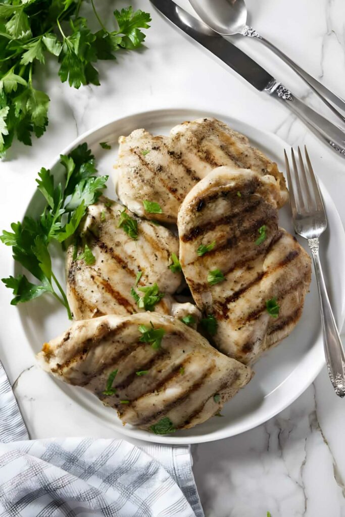 how to grill boneless chicken breasts Four grilled chicken breasts with parsley on a white plate, utensils beside.