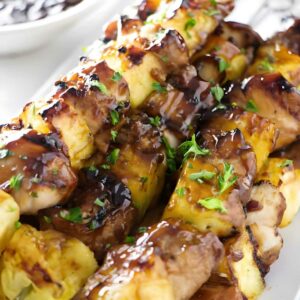Grilled chicken and pineapple skewers with herbs and sauce on a white plate.