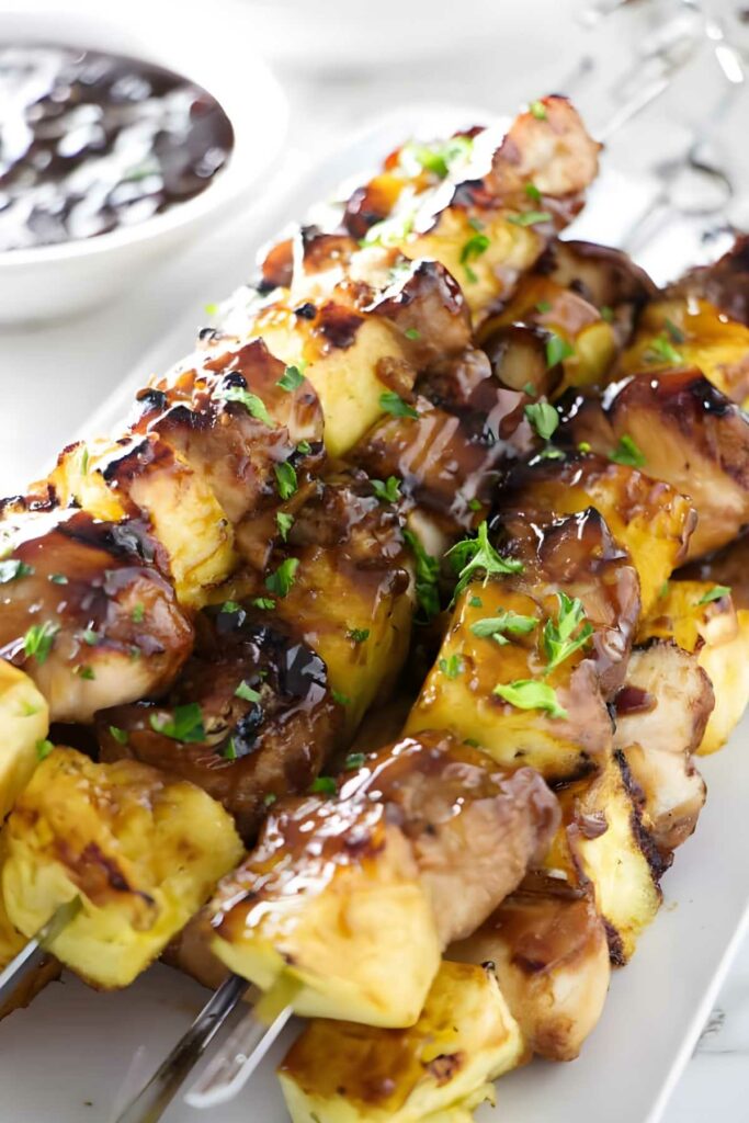 Grilled chicken and pineapple skewers with herbs and sauce on a white plate.