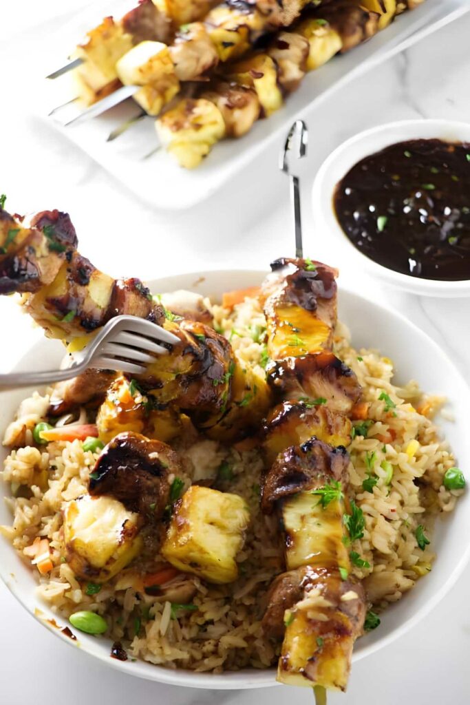 Chicken and pineapple skewers over fried rice in a bowl, fork holding one.