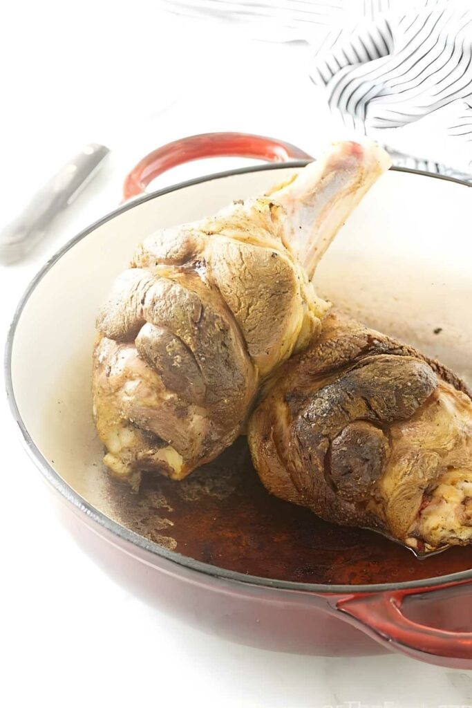 Two browned lamb shanks in a red Dutch oven on a white surface.