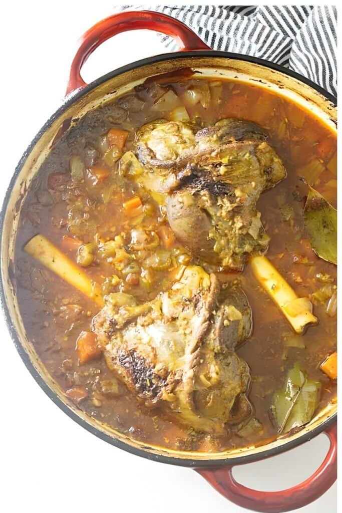 Braised lamb shanks in red Dutch oven with vegetables, viewed from above.