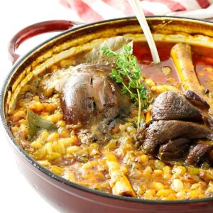 Red Dutch oven with lamb shank stew, chickpeas, veggies, and spoon inside.