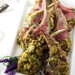 Pistachio crusted rack of lamb on white platter, garnished with herbs.