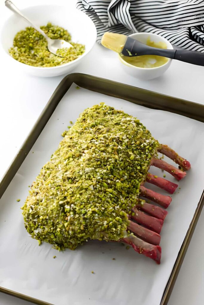 Pistachio-crusted rack of lamb on baking tray with glaze and nuts nearby.