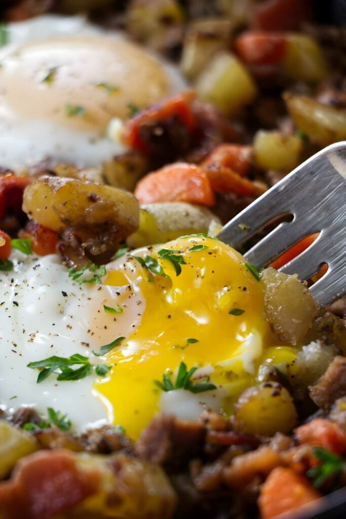 Fork cutting into runny fried egg yolk atop beef hash, close-up view.