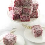 Pink coconut lamington cakes stacked on stand, two plated with forks, white background.