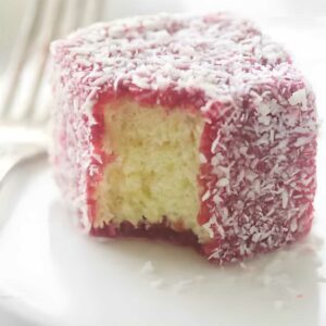 Partially eaten raspberry lamington sponge cake with coconut on a white plate.