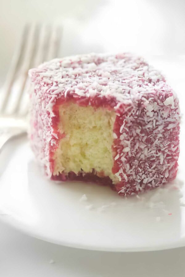 Partially eaten raspberry lamington sponge cake with coconut on a white plate.
