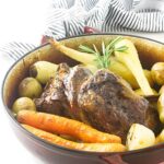 Roasted lamb and vegetables in a red baking dish on white surface.