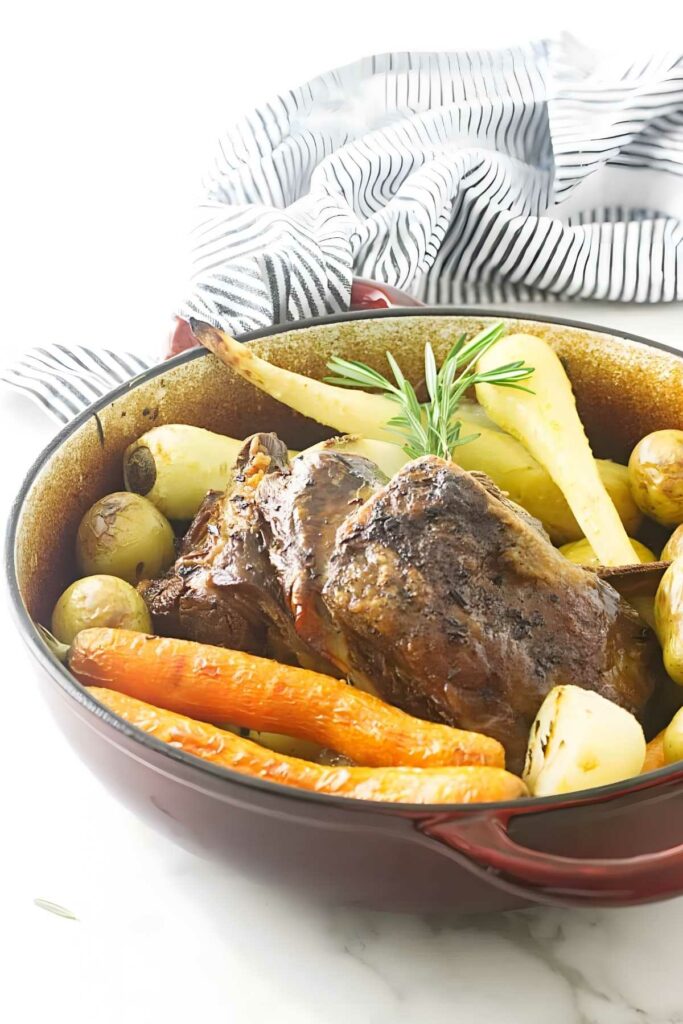Roasted lamb and vegetables in a red baking dish on white surface.