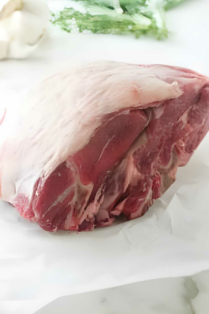 Raw lamb shoulder with white fat on parchment paper, greenery in background.
