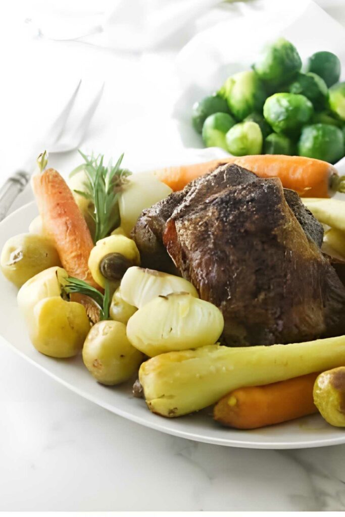 Baked lamb shoulder with vegetables and rosemary on a plate; Brussels sprouts behind.