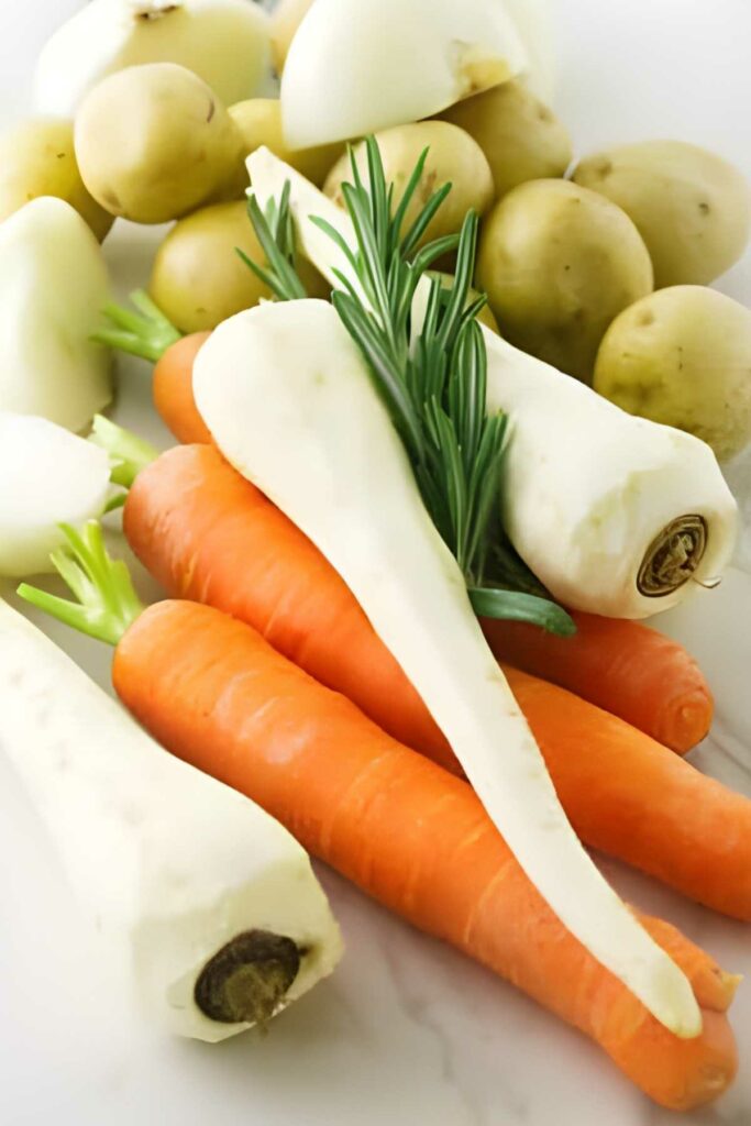 Close-up of raw carrots, parsnips, potatoes, and rosemary on white surface.