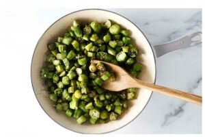 Top-down view of a pan with chopped okra stirred by wooden spoon.