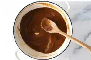 Wooden spoon stirs brown roux in white pot on marble surface.