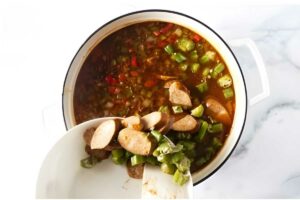 Person adds sausage and green bell pepper to vegetable soup pot.