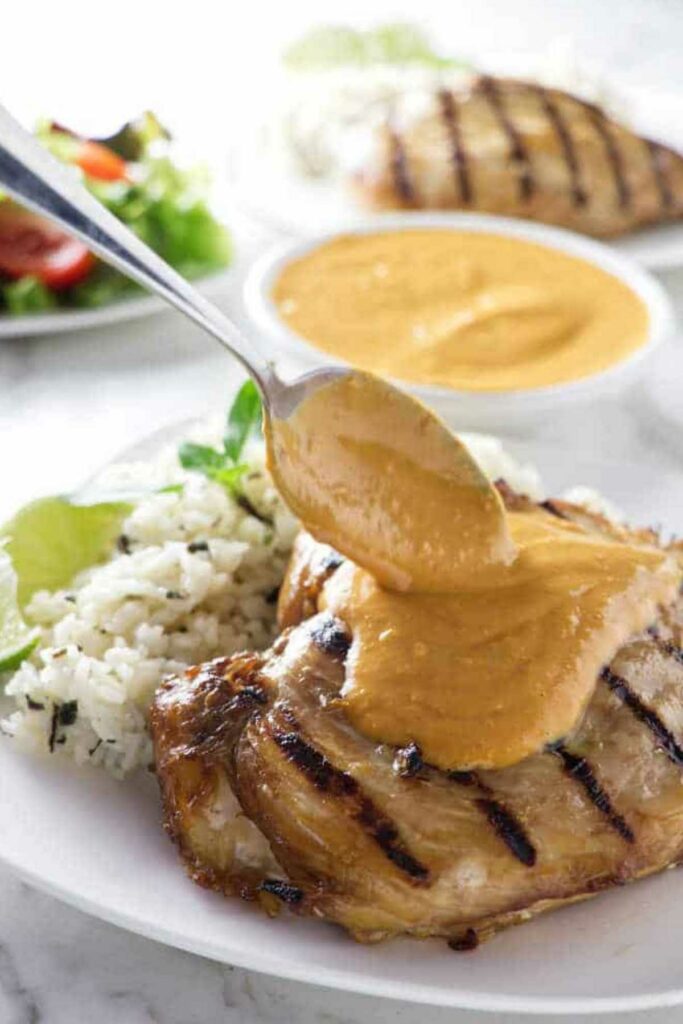 Spoon pours peanut sauce over grilled chicken with rice and lime on plate.