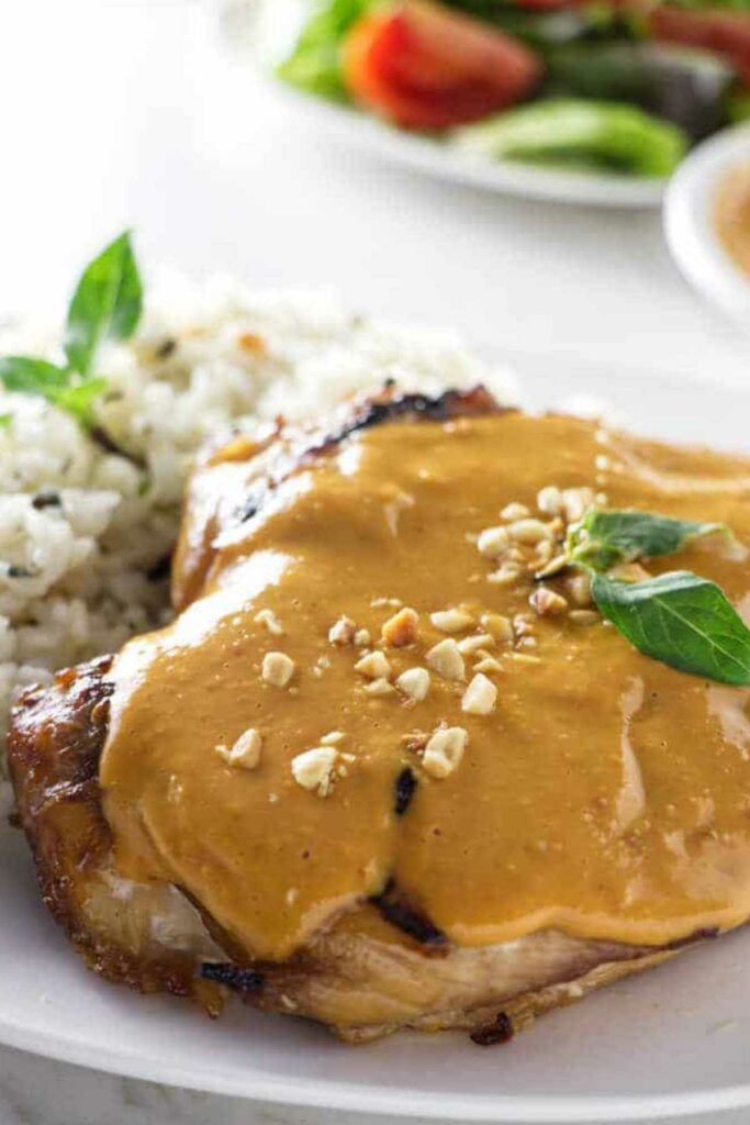 Grilled chicken with peanut sauce, basil, white rice, and salad in background.
