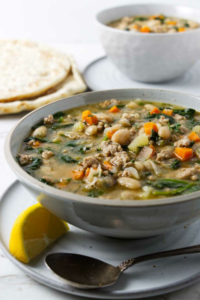 A bowl of white bean soup with chicken and veggies; a lemon wedge next to the soup.