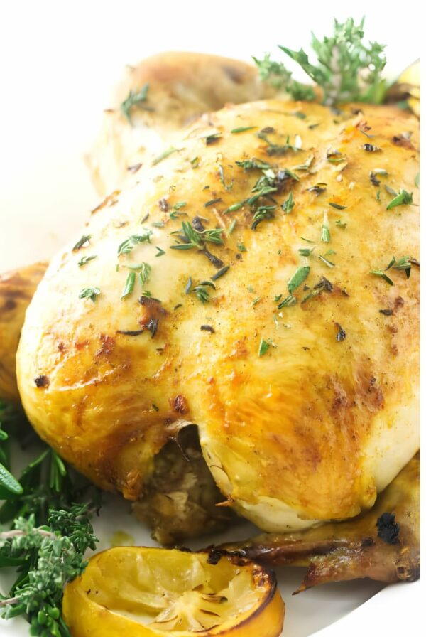 Roasted whole chicken with golden skin, fresh herbs, and lemon slices on plate.
