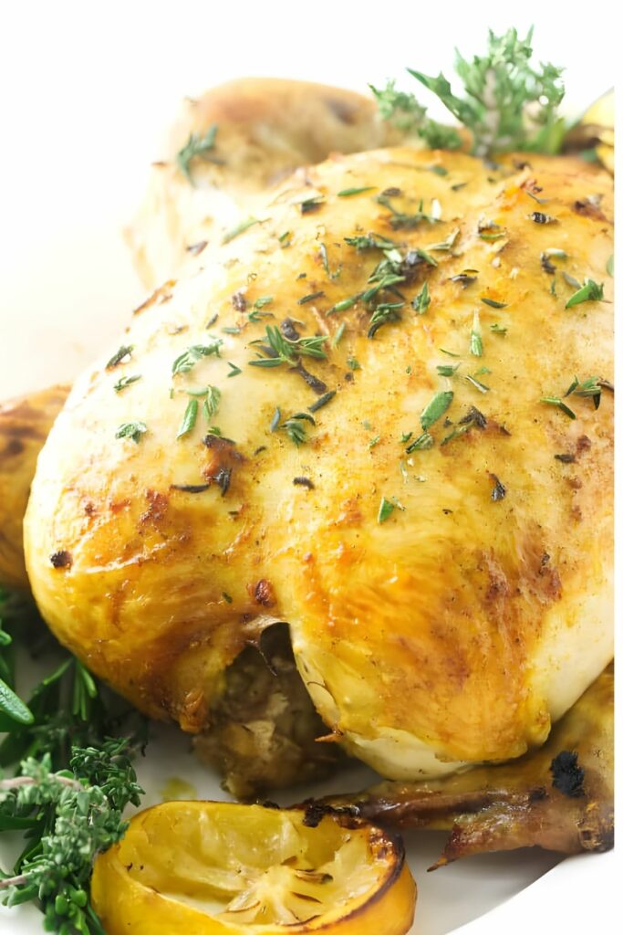 Roasted whole chicken with golden skin, fresh herbs, and lemon slices on plate.