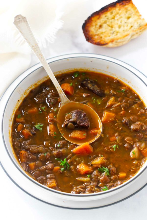A spoon in a bowl of lentil beef soup next to bread.