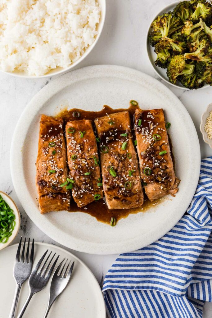 Salmon teriyaki recipe on a plate next to rice and a bowl of broccoli.