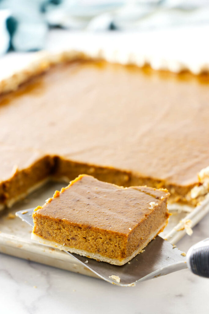 A square slice of pumpkin spice pie in front of a sheet pan with more pie.